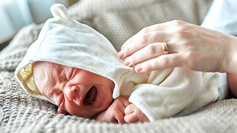 Bebé acostado y llorando mientras una mano acaricia su cara para tranquilizarlo.